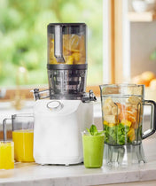 A white juicer on a kitchen counter with glasses of orange and green juice, a blender jar filled with fruit, and a window with greenery in the background.