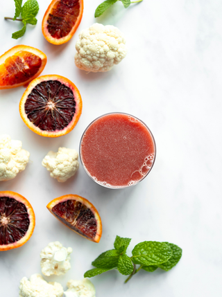 A glass of dark pink minted citrus juice surrounded by sliced blood oranges cauliflower florets and a sprig of mint.