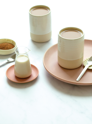 Two ceramic mugs of coffee with a small pitcher of Almond Oat Maple Creamer And a bowl of ground coffee with a spoon