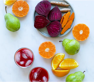two glasses of red juice with ice surrounded by pears oranges and beets