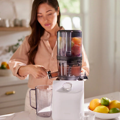 A woman uses the Nama C2, a white multifunction juicer, in a bright kitchen to extract juice from citrus fruits. A clear pitcher and a bowl of lemons and limes are on the counter.