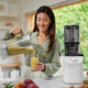A woman in a light green shirt pours fresh juice from a blender pitcher into a glass beside the Nama C2 multifunction juicer on a kitchen counter with fruit, set against a bright window.