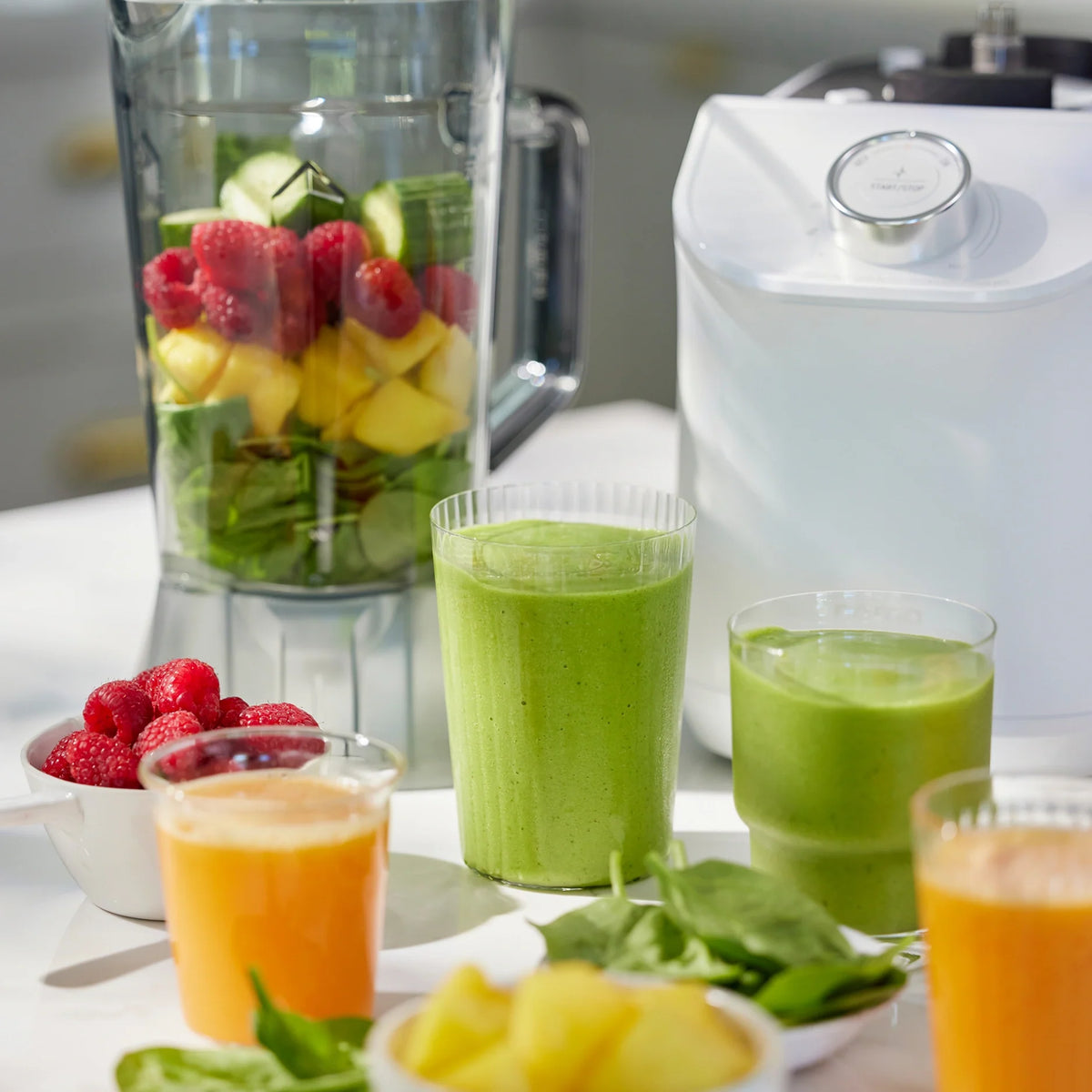 A blender jar filled with spinach, pineapple, raspberries, and cucumber sits next to a white Nama C2 juicer base. Beside it, two green smoothies and two orange juices—freshly made with the multifunctional Nama C2—stand on the counter, surrounded by bowls of raspberries, pineapple, and spinach.