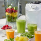 A blender jar filled with spinach, pineapple, raspberries, and cucumber sits next to a white Nama C2 juicer base. Beside it, two green smoothies and two orange juices—freshly made with the multifunctional Nama C2—stand on the counter, surrounded by bowls of raspberries, pineapple, and spinach.