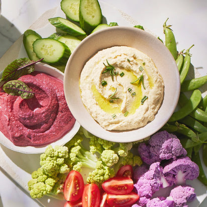 A vibrant plate featuring hummus, beet dip, sliced cucumbers, cherry tomatoes, snap peas, and both green and purple cauliflower shines in natural sunlight—ideal to serve alongside a fresh juice or smoothie made with your Nama C2 multifunction juicer or blender.