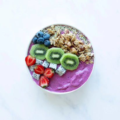 A smoothie bowl with a vibrant purple smoothie base, effortlessly blended using the Nama C2 high-performance blender, topped with sliced kiwi, blueberries, strawberries, dragon fruit cubes, and granola, beautifully arranged on a white marble surface.