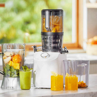 A Nama C2 multifunction juicer and blender sits on a kitchen counter with glasses of green and orange juice, filled with fresh fruit inside the machine, as sunlight streams through a window in the background.