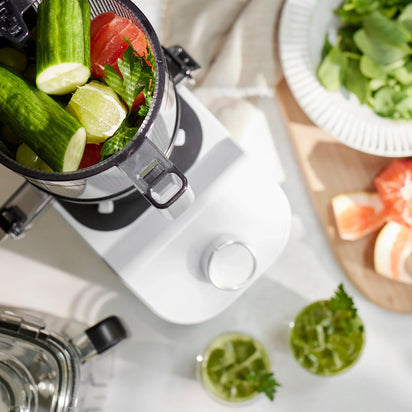 Top view of the Nama C2 cold press juicer filled with cucumbers, grapes, and greens, next to two glasses of green juice, a plate of leafy greens, and grapefruit wedges on a cutting board.