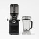 A black Nama C2 cold press juicer with a clear top is placed next to a clear blender pitcher with a lid and handle, both set against a plain white background.
