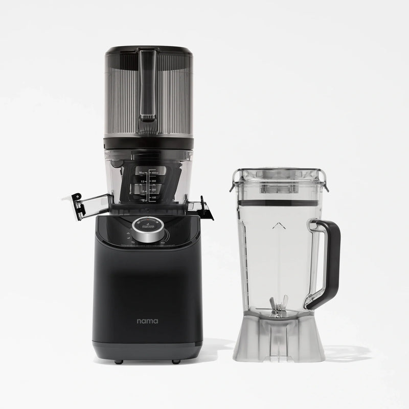 A black Nama C2 cold press juicer with a clear top is placed next to a clear blender pitcher with a lid and handle, both set against a plain white background.