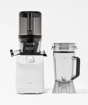 A white Nama juicer with a black top is displayed next to a clear blender pitcher filled with water, both against a plain white background.