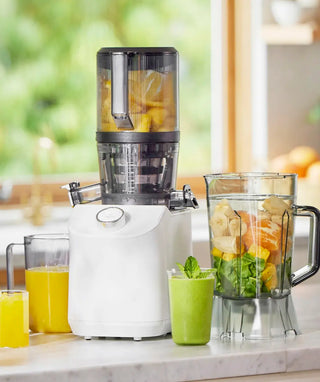 A white juicer on a kitchen counter with glasses of orange and green juice, a blender jar filled with fruit, and a window with greenery in the background.
