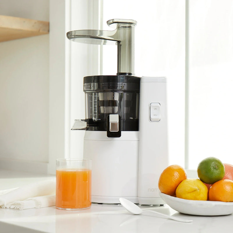 A Nama J1 cold press juicer in white sits on a kitchen counter next to a glass of fresh orange juice, a bowl filled with assorted citrus fruits, a spoon, and a neatly folded white cloth by a bright window—perfect for making nutrient-rich juices every morning.
