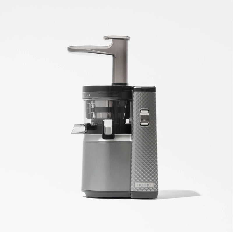 The Nama J1, a modern cold press juicer in silver and black with a transparent container and a textured side panel for enhanced grip, is designed to extract nutrient-rich juices and is displayed against a plain white background.