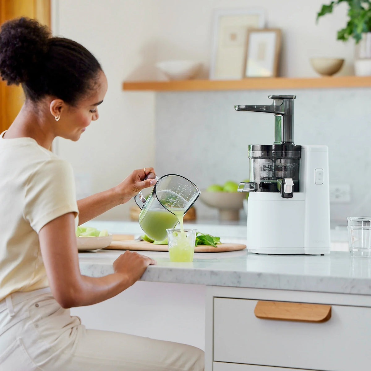 A woman in a light shirt pours fresh, nutrient-rich juice from a pitcher into a glass while sitting at a kitchen counter. The Nama J1 cold press juicer and cut fruits are on the countertop beside her.