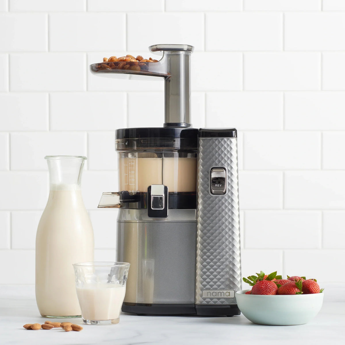 A Nama J1 silver cold press juicer sits on a counter with almond milk inside, accompanied by a glass and carafe of milk, a bowl of strawberries, and a small pile of almonds. The white tiled background highlights how the Nama J1 makes it easy to prepare plant-based milks and nutrient-rich juices at home.
