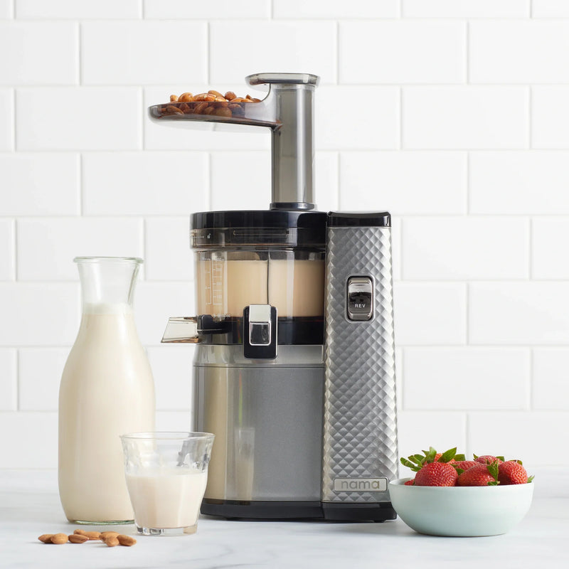 A Nama J1 silver cold press juicer sits on a counter with almond milk inside, accompanied by a glass and carafe of milk, a bowl of strawberries, and a small pile of almonds. The white tiled background highlights how the Nama J1 makes it easy to prepare plant-based milks and nutrient-rich juices at home.
