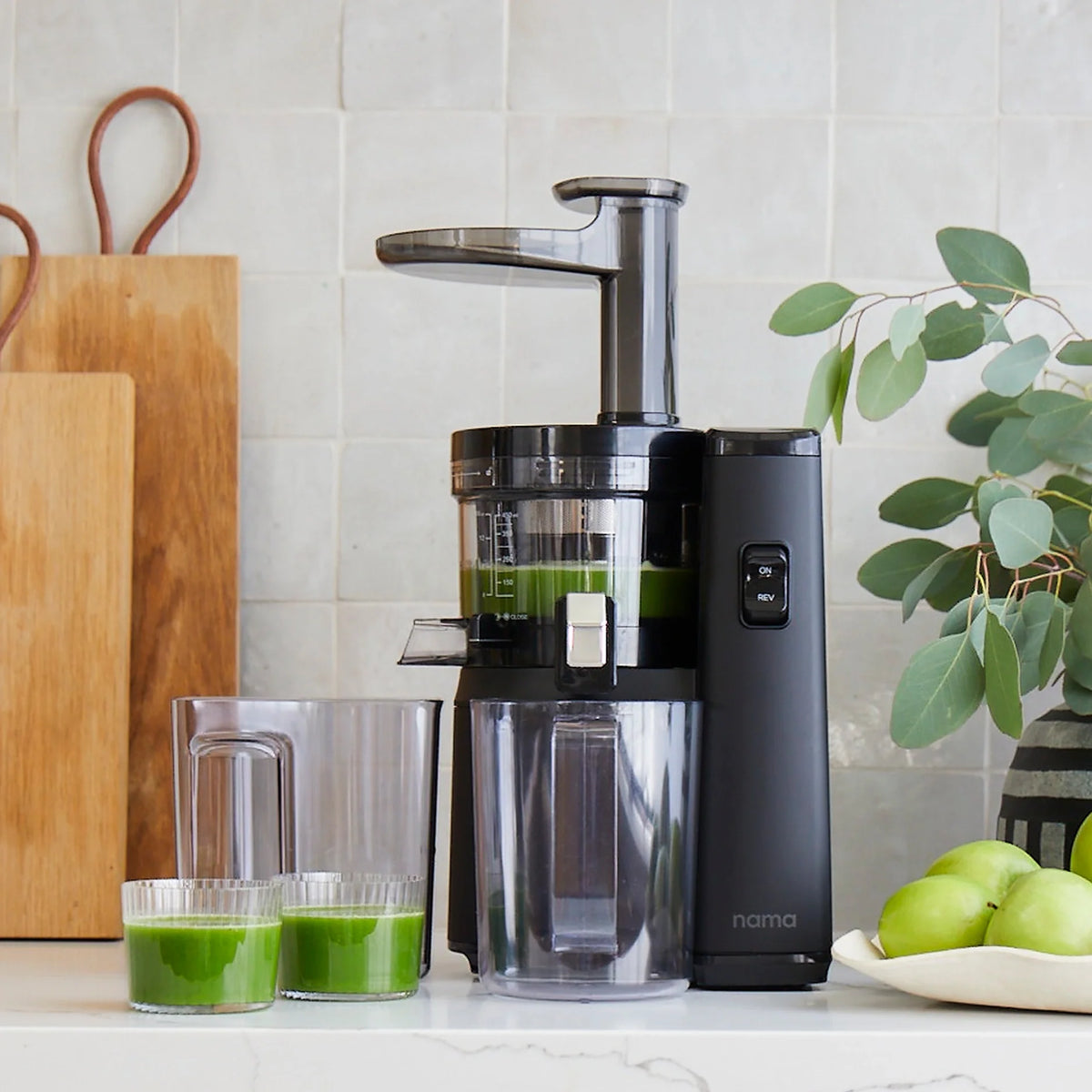 The Nama J1 cold press juicer sits on a kitchen counter, crafting green, nutrient-rich juices into two glasses and a pitcher, surrounded by cutting boards, fresh green apples, and a leafy plant.
