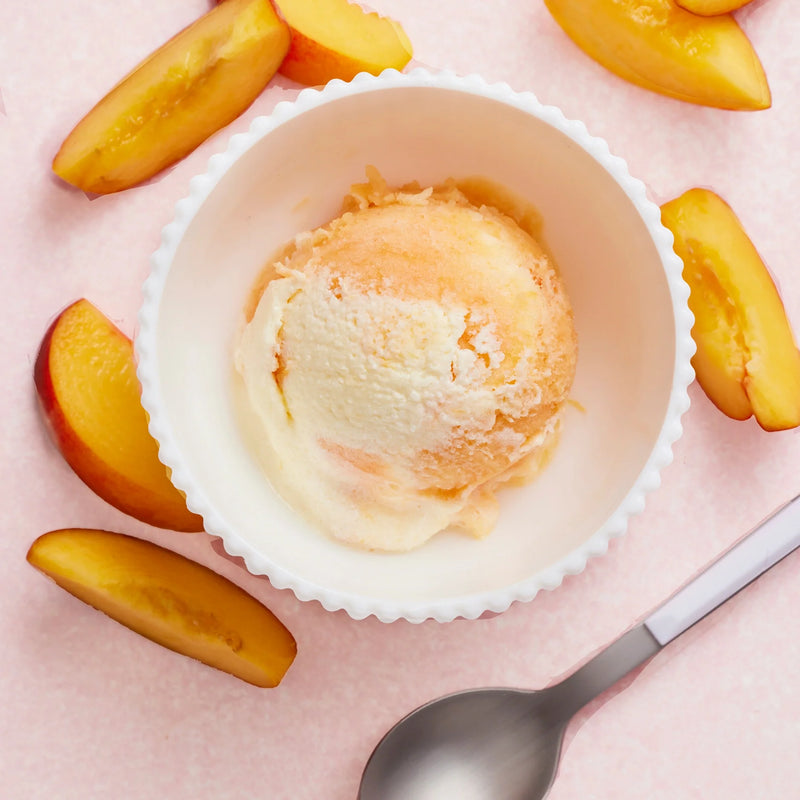 A scoop of Nama J1 peach ice cream, crafted with plant-based milks, sits in a white bowl surrounded by fresh peach slices, with a silver spoon on a light pink surface.