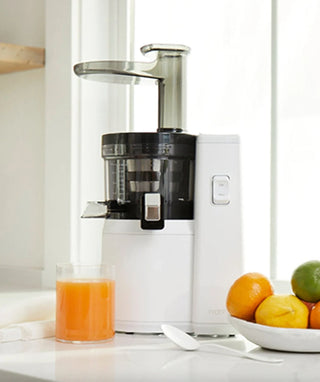 A white juicer sits on a kitchen counter next to a glass of orange juice, a white spoon, and a bowl of assorted citrus fruits by a window.