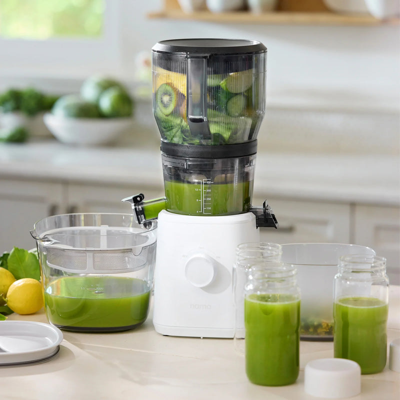 A Nama J2 XL Cold Press Juicer filled with green fruits and vegetables sits on a kitchen counter, dispensing vibrant green juice into glass jars. This easy-to-clean juicer is surrounded by bowls of fresh produce and a lemon, making it perfect for any kitchen routine.