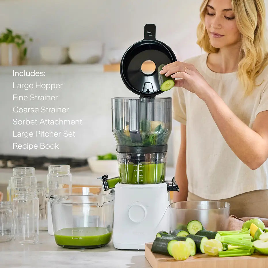 A woman drops a cucumber slice into the Nama J2 XL Cold Press Juicer, as fresh green juice fills the included large pitcher. On-screen text highlights its large hopper, fine and coarse strainers, sorbet attachment, large pitcher set, and recipe book. This easy-to-clean juicer streamlines both preparation and cleanup for your daily juice routine.