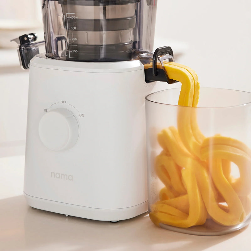 A white Nama J2 XL Cold Press Juicer dispenses thick, yellow juice into a clear glass container. The machine is set to ON and has the brand name nama on the front. The juice flows out in smooth, ribbon-like streams, showcasing the efficiency of this easy-to-clean juicer.