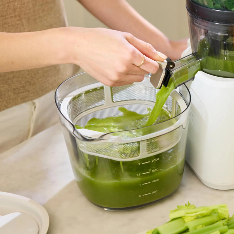 A person uses the Nama J2 XL to extract green juice, pouring it into a clear container with measurement markings. Celery stalks are visible nearby on a countertop, highlighting the benefits of this easy to clean juicer.