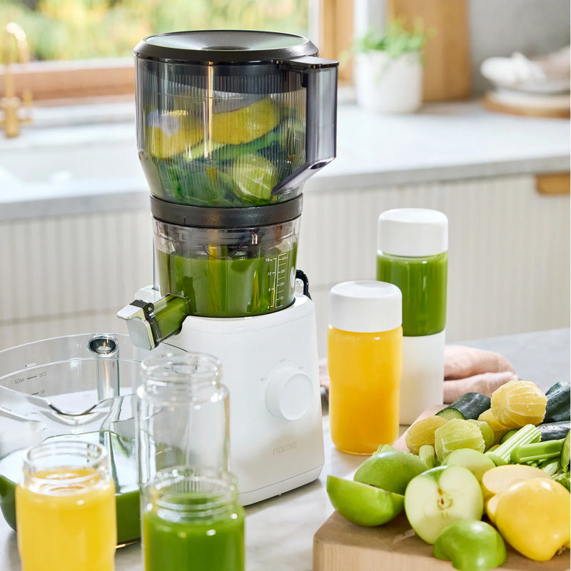 A Nama J2 XL Cold Press Juicer filled with sliced limes and cucumbers sits on a kitchen counter, surrounded by bottles and jars of green and yellow juice, with fresh fruit and vegetables nearby. This cold press juicer is not only efficient but also easy to clean, making your healthy juice preparation both convenient and enjoyable.