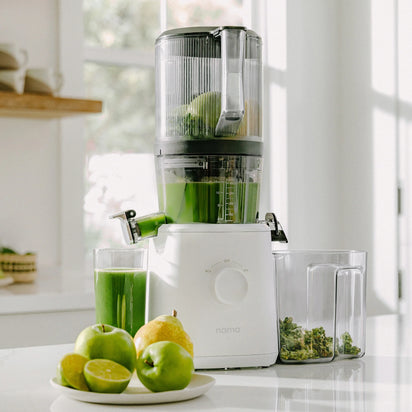 A Nama J2 cold press juicer in white sits on the counter filled with green juice, next to a glass of juice, fresh apples, a lime, and leafy greens in a bright kitchen. This easy-to-clean juicer makes preparing healthy drinks effortless and mess-free.