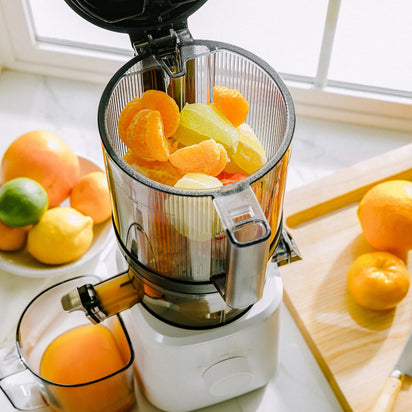 A Nama J2 sits on a kitchen counter, filled with peeled citrus fruits, while whole citrus fruits, a cutting board, and a container of fresh juice are nearby in the sunlight from a window. The Nama J2 is designed for easy cleaning and makes enjoying fresh, homemade juice both effortless and convenient.