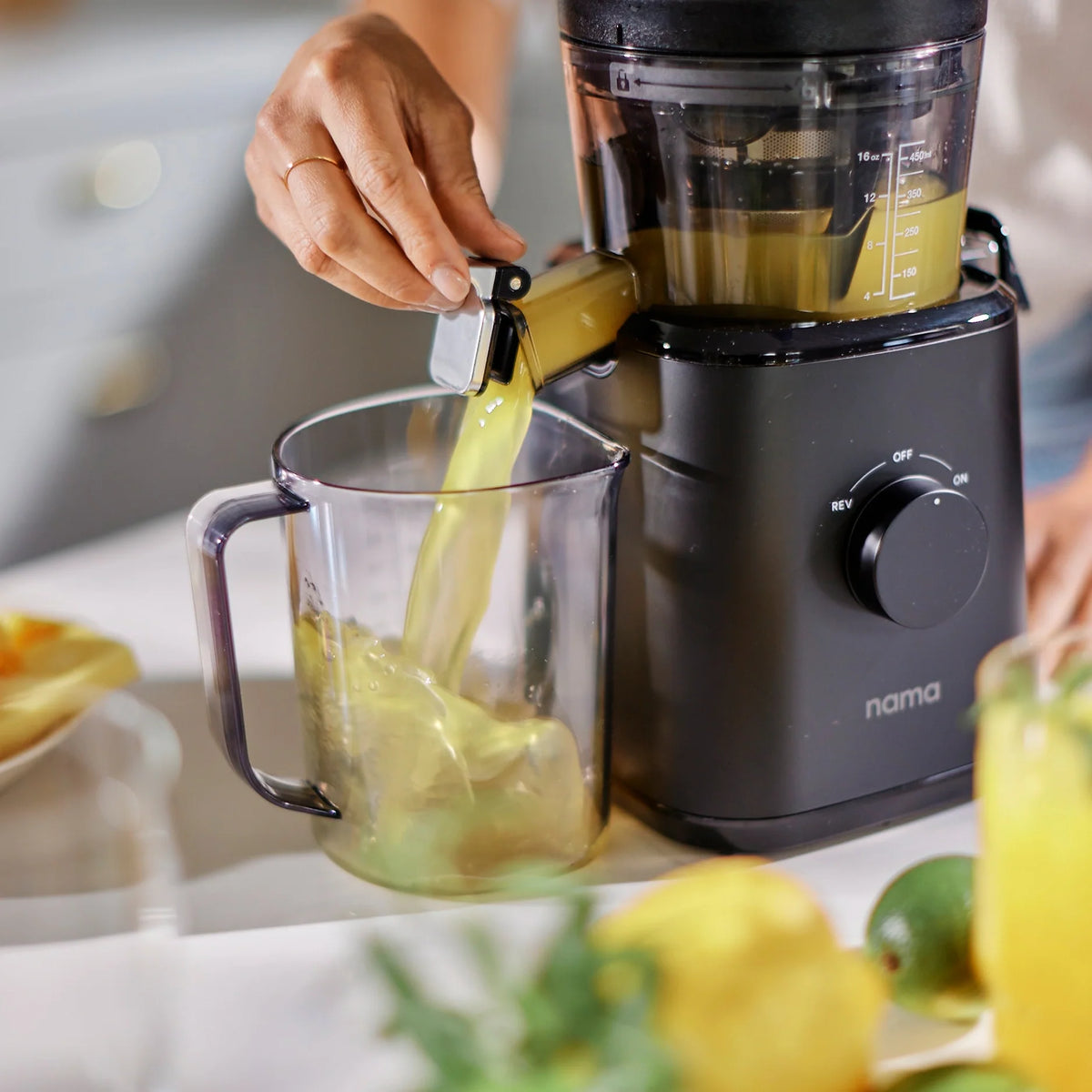 A person pours fresh juice from a black Nama J2 into a clear pitcher on a kitchen counter, with lemons and limes in the foreground. The Nama J2 is easy to clean, making it effortless to prepare delicious, healthy drinks.
