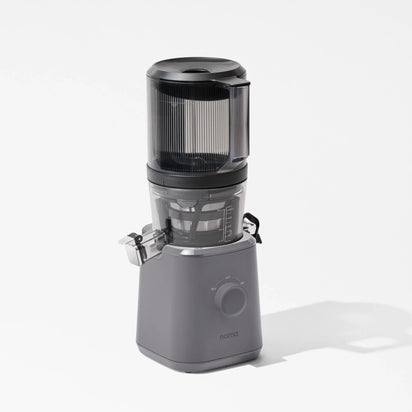 A sleek, gray Nama J2 juicer with a transparent container and lid, featuring a front dial and “nama” branding on the base, is photographed against a white background. Designed for easy cleaning, the Nama J2 is both stylish and practical for everyday cold press juicing.
