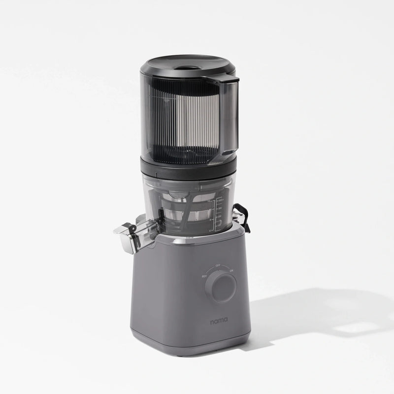 A sleek, gray Nama J2 juicer with a transparent container and lid, featuring a front dial and “nama” branding on the base, is photographed against a white background. Designed for easy cleaning, the Nama J2 is both stylish and practical for everyday cold press juicing.