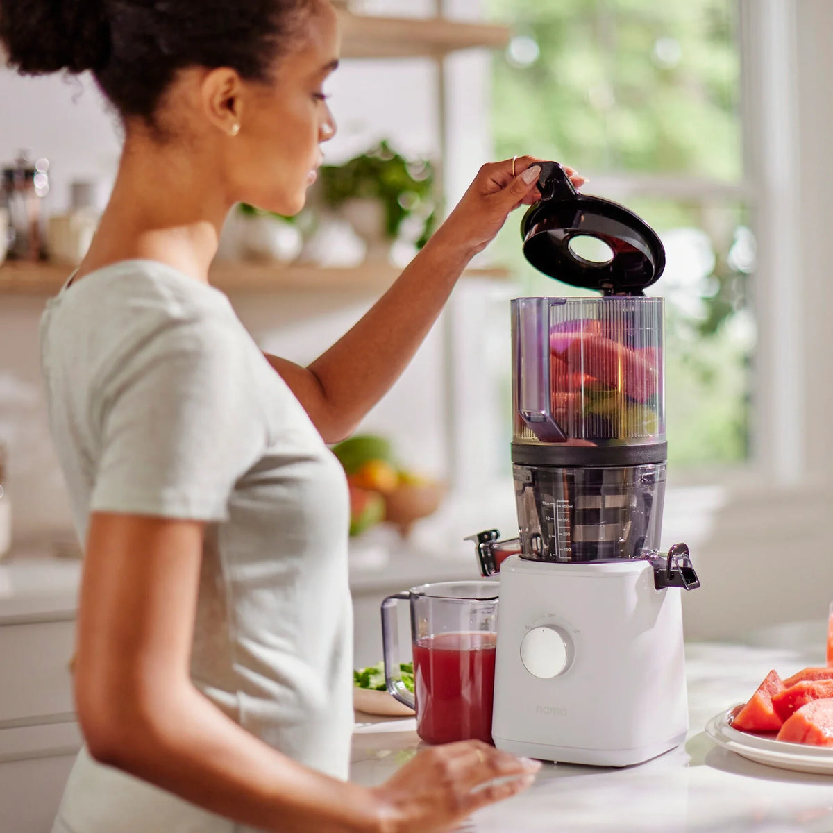 A woman places the lid on a blender filled with fruit in a bright kitchen, where the Nama J2 sits nearby. A glass of juice and cut fruit rest on the counter, showcasing this easy-to-clean juicer as part of her healthy routine.