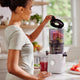 A woman places the lid on a blender filled with fruit in a bright kitchen, where the Nama J2 sits nearby. A glass of juice and cut fruit rest on the counter, showcasing this easy-to-clean juicer as part of her healthy routine.