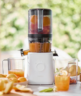 A white juicer with orange juice inside stands on a table with fresh oranges, apricots, juice glasses, and fruit pieces around it. The background is blurred with greenery and sunlight.