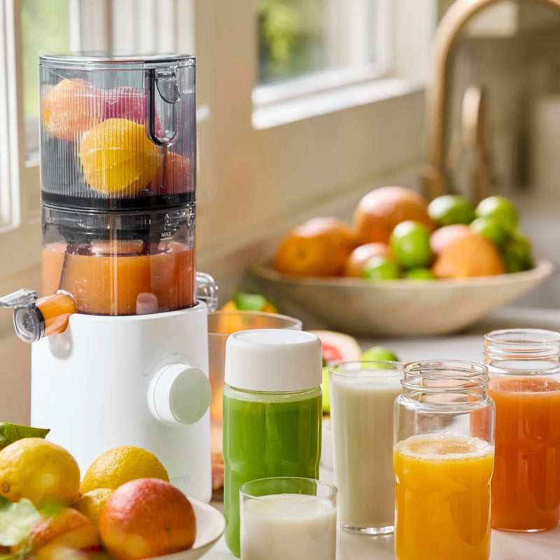 The Nama J3 cold press juicer, filled with citrus fruits, sits on a kitchen counter alongside jars and glasses of fresh juice. In the background, bowls and loose citrus fruits near a sunny window emphasize how easy it is to clean this convenient appliance after preparing your favorite drinks.