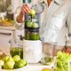 Wearing a light shirt, a person pushes green apples into the white Nama J3 cold press juicer, producing vibrant green juice. Fresh pears, leafy greens, and juice containers are arranged on the counter in a bright kitchen. The Nama J3 is easy to clean and makes juicing simple and efficient.