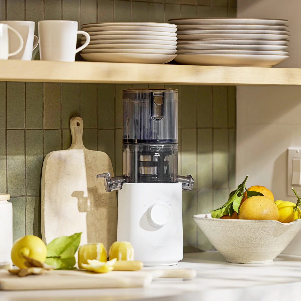A white Nama J3 juicer, known for its easy-to-clean design, sits on a kitchen counter next to a cutting board with cut lemons. Above are shelves stacked with plates and mugs, and a bowl of oranges and lemons is placed beside the juicer.