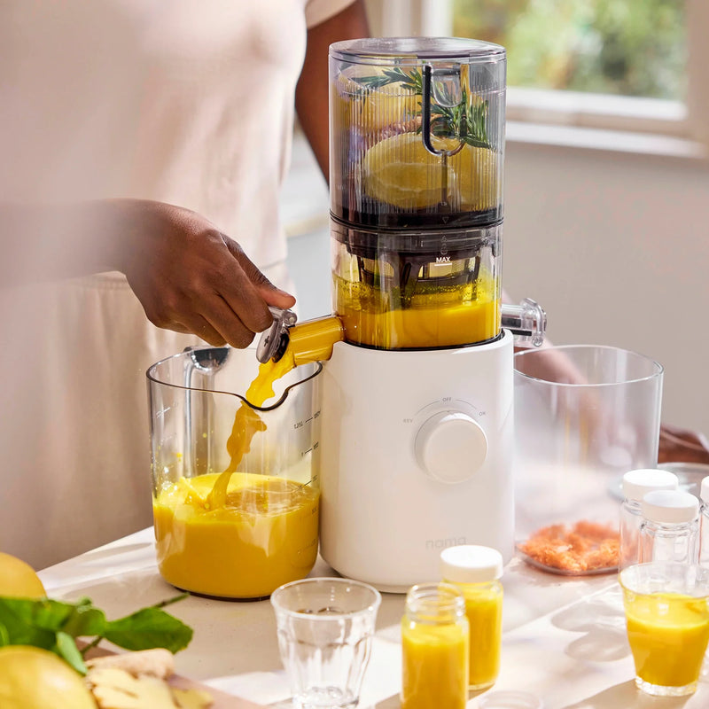 A person uses the Nama J3, a white portable juicer, to make fresh orange juice, pouring it into a clear container. Glasses, juice bottles, fruits, and vegetable pieces are on the counter nearby as sunlight streams through a window. The Nama J3’s easy-to-clean design makes enjoying healthy drinks even simpler.