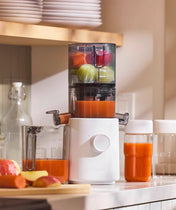 A white juicer on a kitchen counter is making juice from apples and beets. Fresh juice fills two clear containers beside it, with fruit and glass bottles nearby. Sunlight streams onto the countertop.