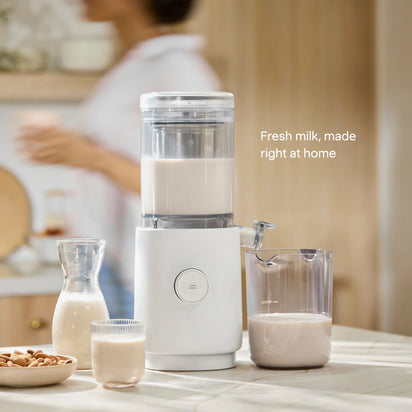 The Nama M1 countertop plant-based milk maker sits alongside a jug, glass, and carafe filled with fresh nut milk. In the background, a person stands blurred. Text reads: Fresh milk—barista-quality made right at home.