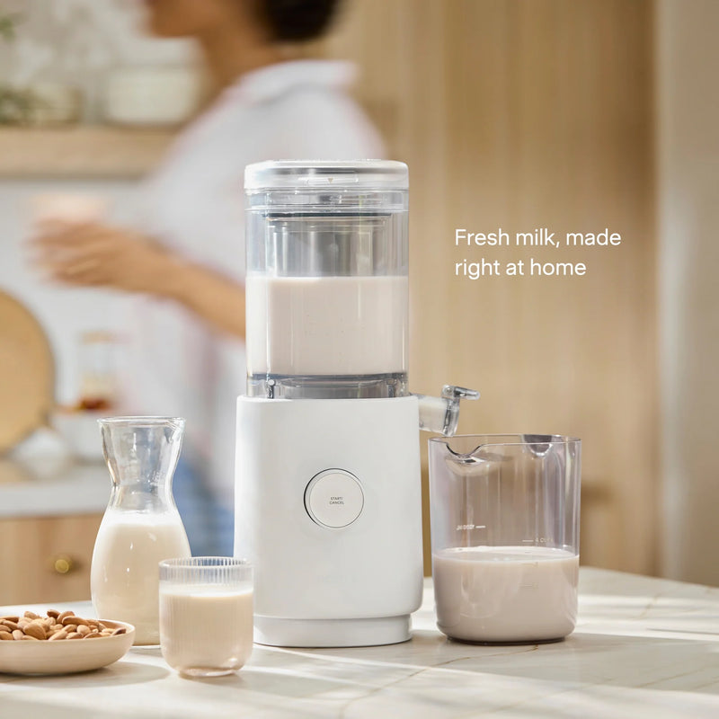 The Nama M1 countertop plant-based milk maker sits alongside a jug, glass, and carafe filled with fresh nut milk. In the background, a person stands blurred. Text reads: Fresh milk—barista-quality made right at home.