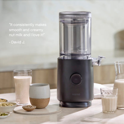 The Nama M1 plant-based milk maker sits on a kitchen counter alongside glasses and a jug of nut milk. A quote on the left reads, “It consistently makes smooth and creamy nut milk and I love it!” – David J. Perfect for creating barista-quality milk at home.