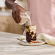 A person pours creamer made with the Nama M1 plant-based milk maker into a glass of iced coffee on a plate with two cinnamon sticks. Text reads, Coffee creamers made your way.