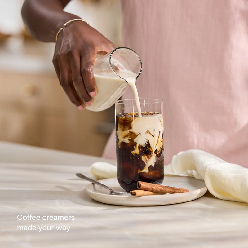 A person pours creamer made with the Nama M1 plant-based milk maker into a glass of iced coffee on a plate with two cinnamon sticks. Text reads, Coffee creamers made your way.