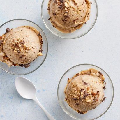 Three glass bowls each hold a scoop of ice cream garnished with chopped nuts and chocolate, showcasing the delicious variety of desserts you can craft with nut milk. A white spoon rests beside the bowls on a light blue surface—perfect for enjoying treats made using your favorite plant-based milk maker, like the Nama M1.