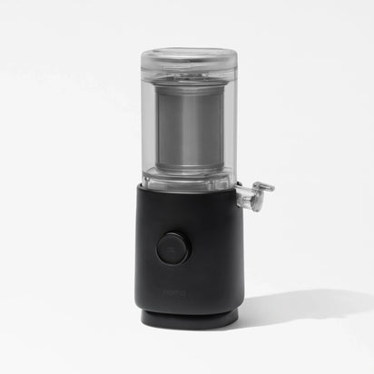 A sleek, black and transparent Nama M1 countertop appliance with a cylindrical glass container on top, a circular control button on the front, and a small side spout is photographed against a white background—perfect for making fresh plant-based nut milk or barista-quality milk at home.