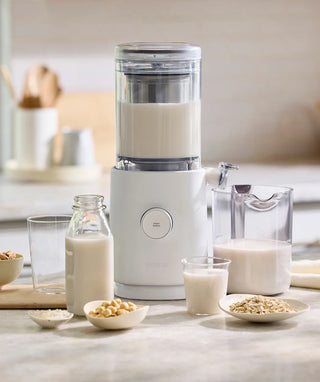 A countertop plant-based milk maker sits among a glass bottle, a pitcher, two glasses filled with milk, and bowls of soybeans, oats, and rice, in a bright kitchen setting.